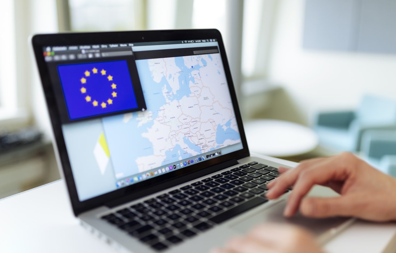 Tietokoneen näytöllä on EU:n lippu ja Euroopan kartta. Oikea käsi koskettaa kosketushiirtä.