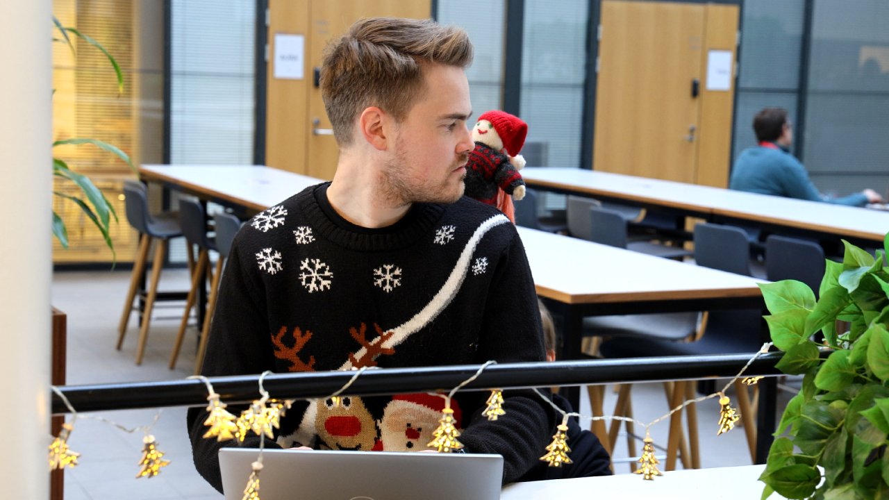 Kuvan keskellä asiantuntija käyttää kannettavaa tietokonetta ja hänellä on musta jouluvillapaita, jossa on poron ja tontun kuva. Hän on kääntynyt katsomaan vasemmalle olkapäälleen, sillä sen takaa kurkistelee tonttunukke.