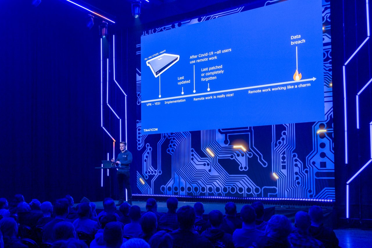 operaatiopäällikkö Matias Mesiä sinihohteisten piirilevykoristeiden ympäröimänä Disobeyn esiintymislavalla , taustalla esitelmäkalvolla aikajana, joka kuvaa sitä, miten hybridityöhön siirtyminen ja unohdettu reititin saattavat johtaa vuosien aikajänteellä tietomurtoon.