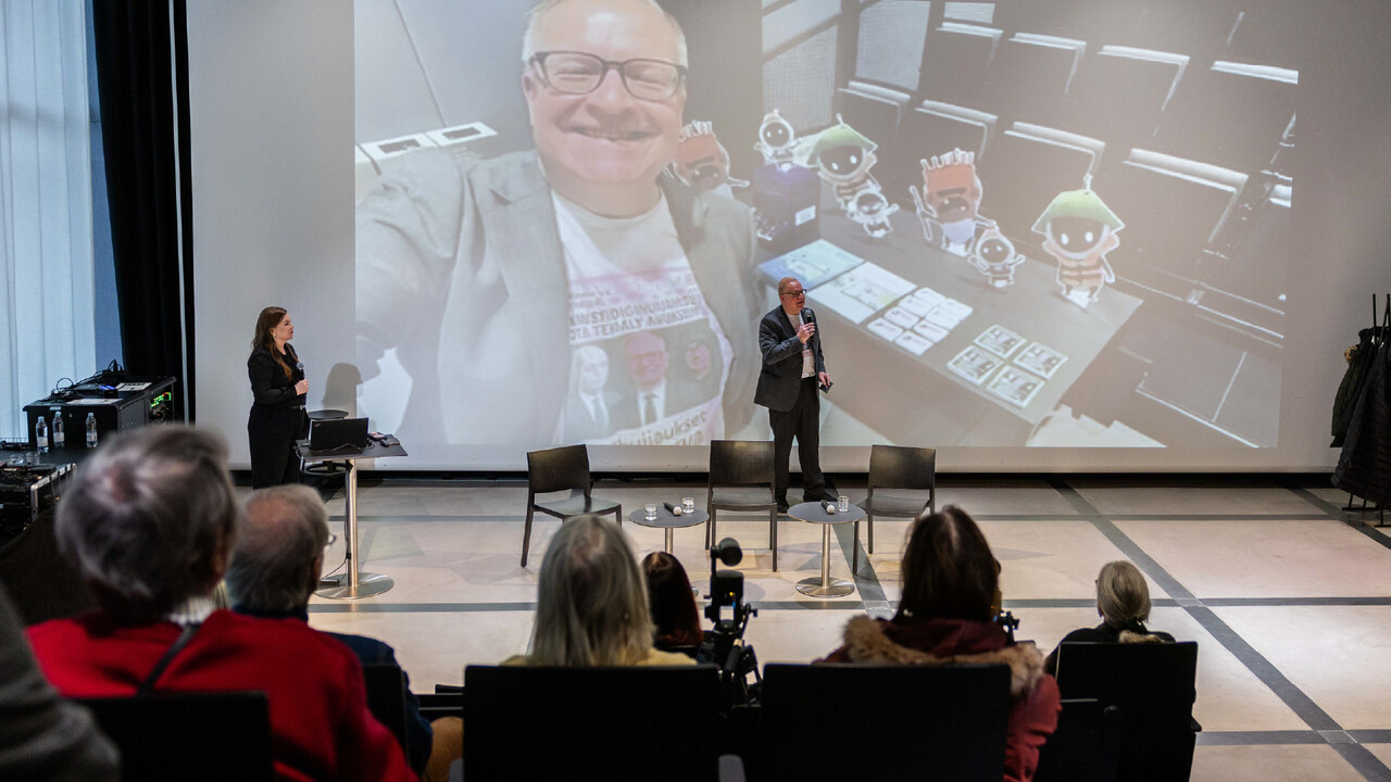 Digi- ja väestötietoviraston johtava erityisasiantuntija Kimmo Rousku Oodissa esiintymislavalla, taustalla pyörii tekoälyllä manipuloitu Rouskun kuvaama selfie-video.